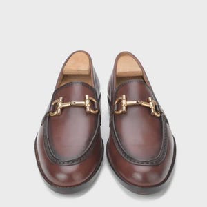 Puede incluir: Un par de mocasines de cuero marr&oacute;n con un detalle de adorno de metal dorado. Los zapatos tienen un dise&ntilde;o cl&aacute;sico con punta redondeada y un ribete marr&oacute;n oscuro alrededor de la suela. Los mocasines se muestran sobre un fondo blanco.