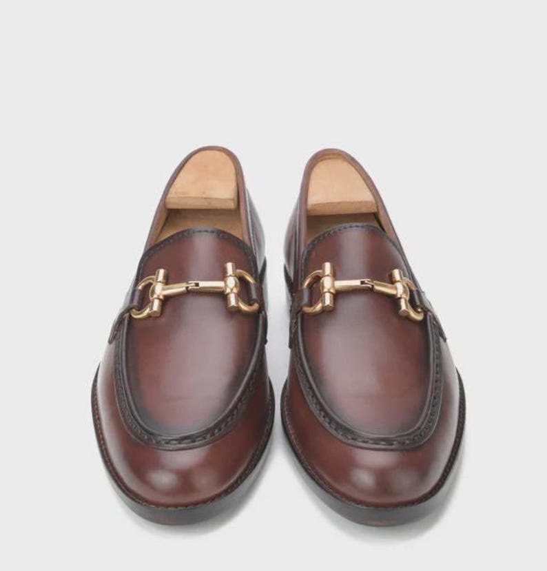 Puede incluir: Un par de mocasines de cuero marr&oacute;n con un detalle de adorno de metal dorado. Los zapatos tienen un dise&ntilde;o cl&aacute;sico con punta redondeada y un ribete marr&oacute;n oscuro alrededor de la suela. Los mocasines se muestran sobre un fondo blanco.