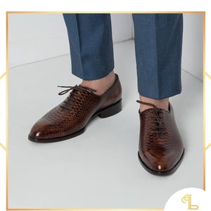 Op de afbeelding: Bruine leren geklede schoenen met een textuur slangenpatroon en vetersluiting. De schoenen hebben een gepolijste afwerking en een lage hak. De schoenen worden gedragen met een blauwe broek.