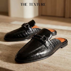 Peut inclure: Une paire de mules noires avec une texture en cuir gaufré façon crocodile brillant et un mors décoratif en métal noir sur le dessus. La semelle intérieure est de couleur beige et le texte THE TEXTURE est visible en haut de l'image.