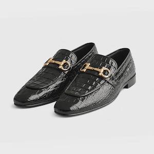 Puede incluir: Un par de mocasines de cuero negro con textura de cocodrilo. Cada zapato presenta una hebilla dorada en la parte superior. Los zapatos tienen un acabado brillante y un diseño clásico y formal.