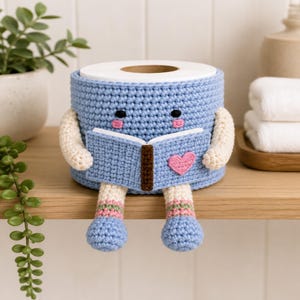 Könnte beinhalten: Ein hellblauer, gehäkelter Toilettenpapierüberzug in Form einer Figur, die ein Buch liest. Die Figur hat cremefarbene Arme und Beine, rosa Wangen und ein kleines rosa Herz auf dem blauen Buchdeckel. Das Design zeigt einen braunen Buchrücken.