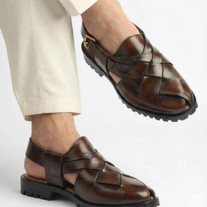 May include: Brown leather sandals with a woven design. The sandals have a strap around the ankle and a black sole. The shoes are shown on a person wearing cream-colored pants.