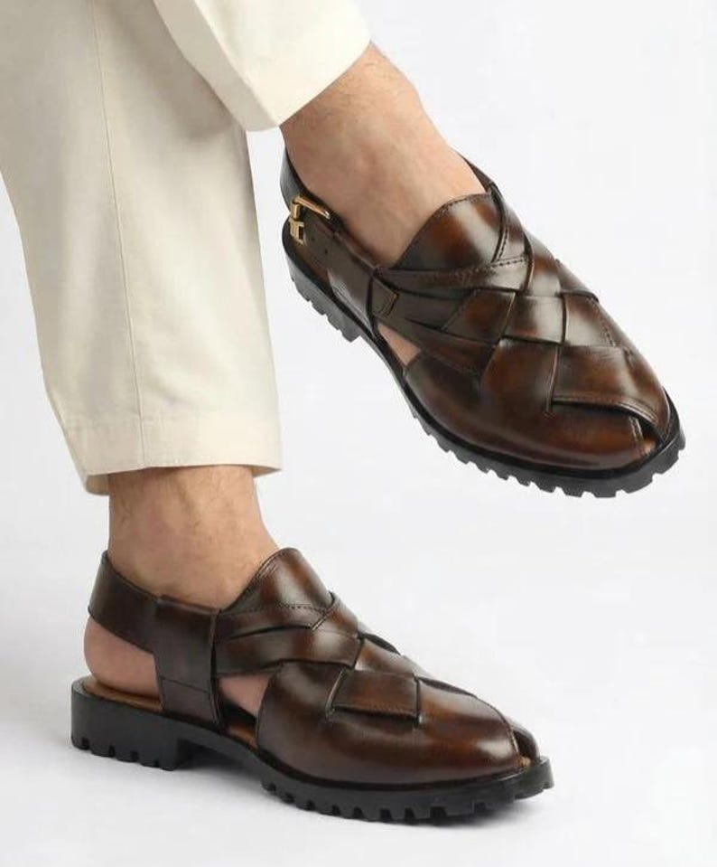 May include: Brown leather sandals with a woven design. The sandals have a strap around the ankle and a black sole. The shoes are shown on a person wearing cream-colored pants.