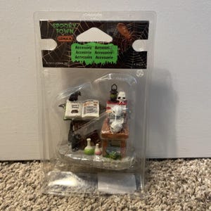 May include: A Lemax Spooky Town miniature accessory set in clear plastic packaging. The set includes a small wooden table with stacked books, three white skulls, a black lantern, an open book with a black crow, and two small glass bottles.