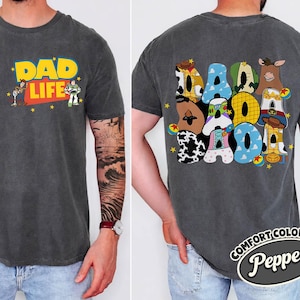 May include: Dark gray t-shirt with "DAD LIFE" in yellow and red, featuring cartoon characters. The back of the shirt displays "DAD" in colorful, character-themed letters. The shirt is labeled "Comfort Colors Pepper".