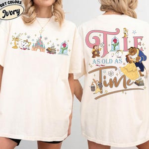Può includere: T-shirt color avorio con grafica colorata de La Bella e la Bestia. Il davanti presenta personaggi e un castello. Il retro mostra le parole "Tale as Old as Time" con Belle e la Bestia.