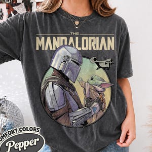 Puede incluir: Una camiseta de manga corta gris carb&oacute;n con un estampado gr&aacute;fico de The Mandalorian y Grogu dentro de un marco circular. El texto THE MANDALORIAN est&aacute; impreso en letras beige grandes sobre la ilustraci&oacute;n, que tambi&eacute;n incluye una nave espacial.