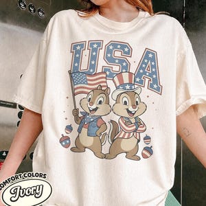 May include: Ivory-colored t-shirt featuring the word "USA" in blue with star accents. Two cartoon chipmunks are dressed in patriotic attire, one holding an American flag. Decorative red, white, and blue ornaments are scattered around the chipmunks.