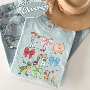 May include: A light blue t-shirt featuring a graphic print of various cartoon characters and colorful bows. The shirt is displayed alongside a pair of light wash denim jeans, a tan wide-brimmed hat, and a silver necklace. Text on the shirt reads Comfort Colors Chambray.
