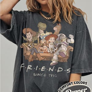 Puede incluir: Camiseta gris oscuro de gran tama&ntilde;o con un gr&aacute;fico de personajes de Toy Story en un sof&aacute;, con la palabra "FRIENDS" y "SINCE 1995" impresas debajo. La camiseta tiene una etiqueta "Comfort Colors Pepper".