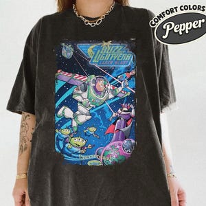 Pode incluir: Uma t-shirt preto carv&atilde;o com uma estampa gr&aacute;fica de Buzz Lightyear, Zurg e alien&iacute;genas verdes no espa&ccedil;o. O texto diz Buzz Lightyear Laser Blast e Disneyland. Um log&oacute;tipo Comfort Colors Pepper &eacute; vis&iacute;vel no canto superior direito.