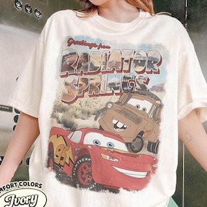 May include: Ivory-colored t-shirt featuring a graphic of Lightning McQueen and Mater from the movie Cars, with the text "Greetings from Radiator Springs." The shirt is from Comfort Colors.