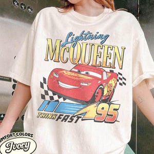 K&ouml;nnte beinhalten: Elfenbeinfarbenes T-Shirt mit einem Aufdruck von Lightning McQueen aus dem Film Cars. Das Design enth&auml;lt den Text "Lightning McQueen", "Think Fast" und die Nummer "95". Das Shirt ist von der Marke Comfort Colors.