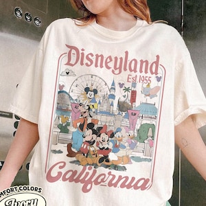 Może przedstawiać: Kremowa koszulka z grafiką Disneyland. Projekt przedstawia Myszka Miki, Minnie, Kaczora Donalda i inne postacie Disneya przed diabelskim młynem i zamkiem. Tekst "Disneyland Est. 1955 California" jest wydrukowany w r&oacute;żowo-czerwonym kolorze.