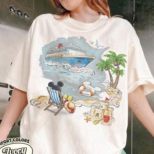 Op de afbeelding: Ivoor kleurig T-shirt met een kleurrijke strandsc&egrave;ne met een cruiseschip, palmbomen en Mickey Mouse. Het shirt heeft de woorden "Comfort Colors" en "Ivory" erop gedrukt.