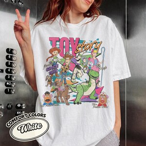 May include: White t-shirt featuring a colorful graphic of Toy Story characters, including Woody, Buzz Lightyear, and Jessie. The design includes the text "TOY STORY" and "Since 1995". The shirt also has the Comfort Colors logo.