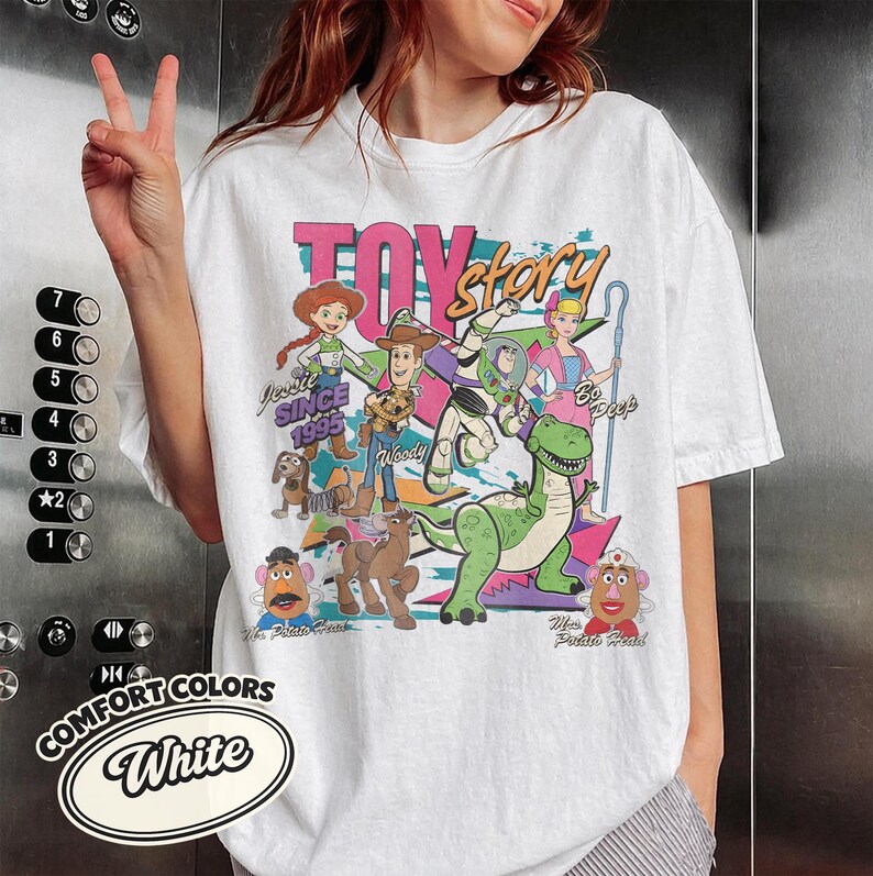 May include: White t-shirt featuring a colorful graphic of Toy Story characters, including Woody, Buzz Lightyear, and Jessie. The design includes the text "TOY STORY" and "Since 1995". The shirt also has the Comfort Colors logo.