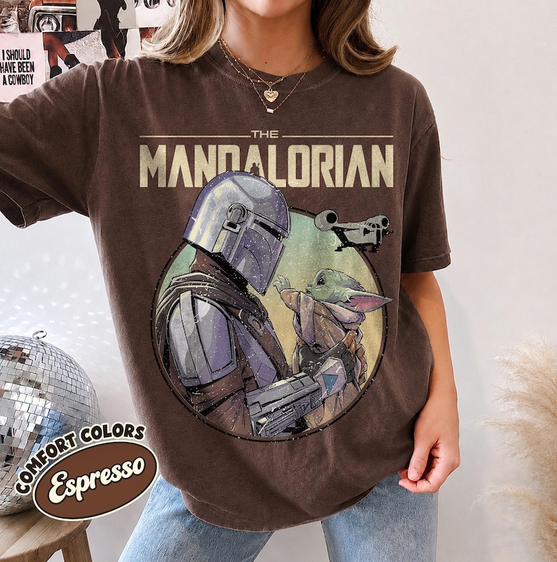 Puede incluir: Una camiseta marr&oacute;n de manga corta con un estampado gr&aacute;fico de The Mandalorian y Grogu dentro de un marco circular. El texto THE MANDALORIAN est&aacute; impreso en letras beige grandes sobre el gr&aacute;fico, con una peque&ntilde;a silueta de nave espacial.