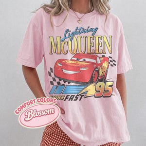 K&ouml;nnte beinhalten: Hellrosa T-Shirt mit einem Aufdruck von Lightning McQueen aus dem Film Cars. Das Design enth&auml;lt den Text "Lightning McQueen" &uuml;ber einem roten Rennwagen mit der Nummer "95". Das Shirt hat auch die Worte "Think Fast".
