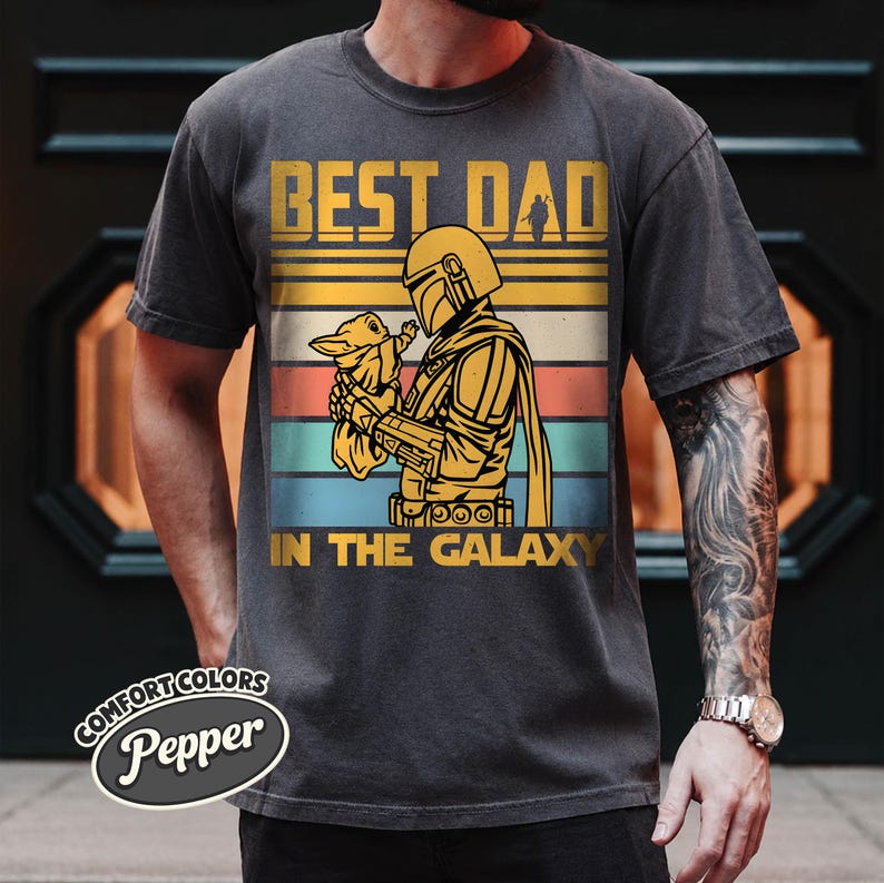 以下が含まれることがあります： 「BEST DAD IN THE GALAXY」の文字と、小さな生き物を抱えるマンダロリアンキャラクターのグラフィックがプリントされたダークグレーのTシャツ。レトロな配色と水平ストライプのデザインが特徴です。