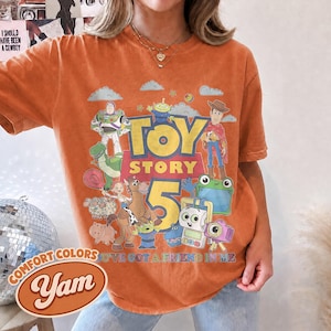 May include: An orange short-sleeve t-shirt featuring a graphic print of Toy Story characters including Buzz Lightyear, Woody, and Rex. The text reads Toy Story 5 and You've Got A Friend In Me. The shirt has a crew neckline and a relaxed fit.