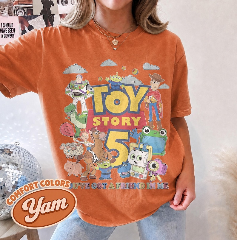 May include: An orange short-sleeve t-shirt featuring a graphic print of Toy Story characters including Buzz Lightyear, Woody, and Rex. The text reads Toy Story 5 and You've Got A Friend In Me. The shirt has a crew neckline and a relaxed fit.