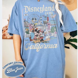 Może przedstawiać: Niebieska koszulka w kolorze dżinsu z grafiką Disneyland. Projekt zawiera kultowe postacie, takie jak Myszka Miki i Myszka Minnie, Kaczor Donald, a także logo Disneyland i napis "California". Koszulka marki Comfort Colors.
