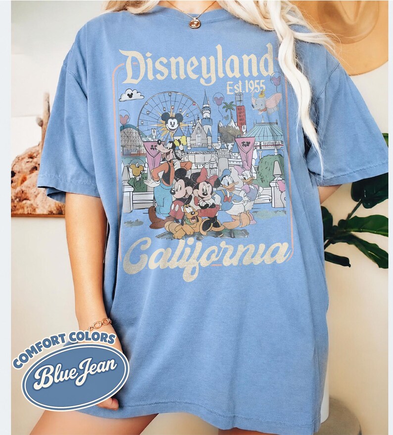 Może przedstawiać: Niebieska koszulka w kolorze dżinsu z grafiką Disneyland. Projekt zawiera kultowe postacie, takie jak Myszka Miki i Myszka Minnie, Kaczor Donald, a także logo Disneyland i napis "California". Koszulka marki Comfort Colors.