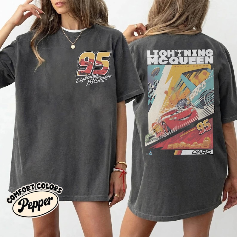 May include: Dark gray t-shirt featuring the number 95 and "Lightning McQueen" text on the front. The back of the shirt displays a colorful racing scene with "Lightning McQueen" and "95" prominently featured. The shirt is from the "Cars" movie.