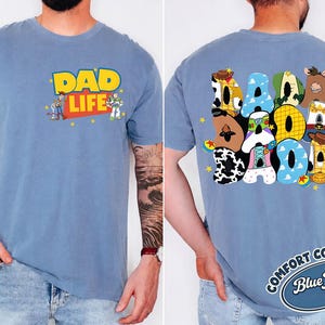 May include: A blue jean-colored t-shirt with the words "DAD LIFE" in yellow and red, featuring cartoon characters. The back of the shirt displays the word "DAD" in colorful, cartoon-themed letters. The shirt is a Comfort Colors brand.