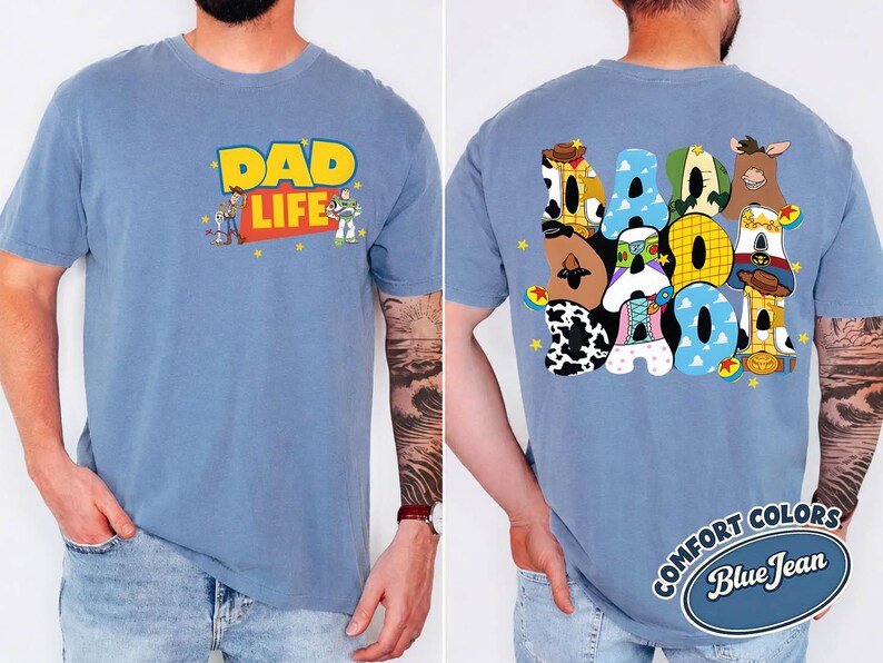 May include: A blue jean-colored t-shirt with the words "DAD LIFE" in yellow and red, featuring cartoon characters. The back of the shirt displays the word "DAD" in colorful, cartoon-themed letters. The shirt is a Comfort Colors brand.