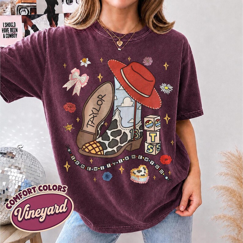 Peut inclure: Un t-shirt &agrave; manches courtes bordeaux fonc&eacute; avec une impression graphique repr&eacute;sentant une botte de cow-boy &agrave; taches de vache, un chapeau rouge, un n&oelig;ud rose et des blocs avec les lettres T, S et 13. Le texte Taylor et la phrase I KNEW IT I KNEW YOU.