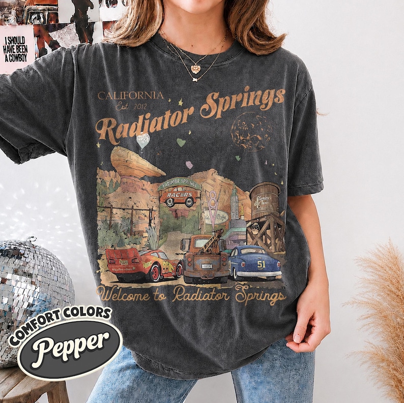 May include: A charcoal grey short-sleeve t-shirt featuring a graphic print of Radiator Springs with the text California Est. 2017, Radiator Springs, and Welcome to Radiator Springs. The design includes a red race car, a brown tow truck, and a blue car.