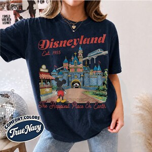 May include: A dark navy blue t-shirt featuring a graphic print of a castle, a monorail, and a cartoon mouse. The text reads Disneyland Est. 1955 and The Happiest Place On Earth. A small circular label in the corner displays the text Comfort Colors True Navy.