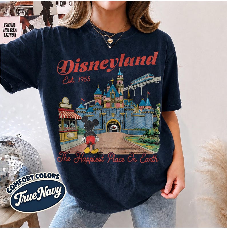 May include: A dark navy blue t-shirt featuring a graphic print of a castle, a monorail, and a cartoon mouse. The text reads Disneyland Est. 1955 and The Happiest Place On Earth. A small circular label in the corner displays the text Comfort Colors True Navy.