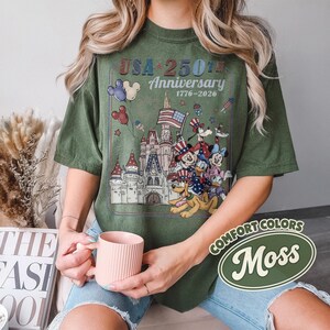 Puede incluir: Camiseta verde oliva con un gr&aacute;fico que celebra el 250 aniversario de EE. UU. El dise&ntilde;o incluye un castillo, personajes de dibujos animados y el texto "USA 250th Anniversary 1776-2026". La camiseta est&aacute; etiquetada como "Comfort Colors Moss".
