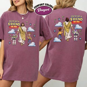 May include: A maroon Comfort Colors t-shirt with a graphic of Jesus and cartoon characters. The text "YOU'VE GOT A FRIEND IN ME" is printed on the front and back. The shirt has a crew neck and short sleeves.