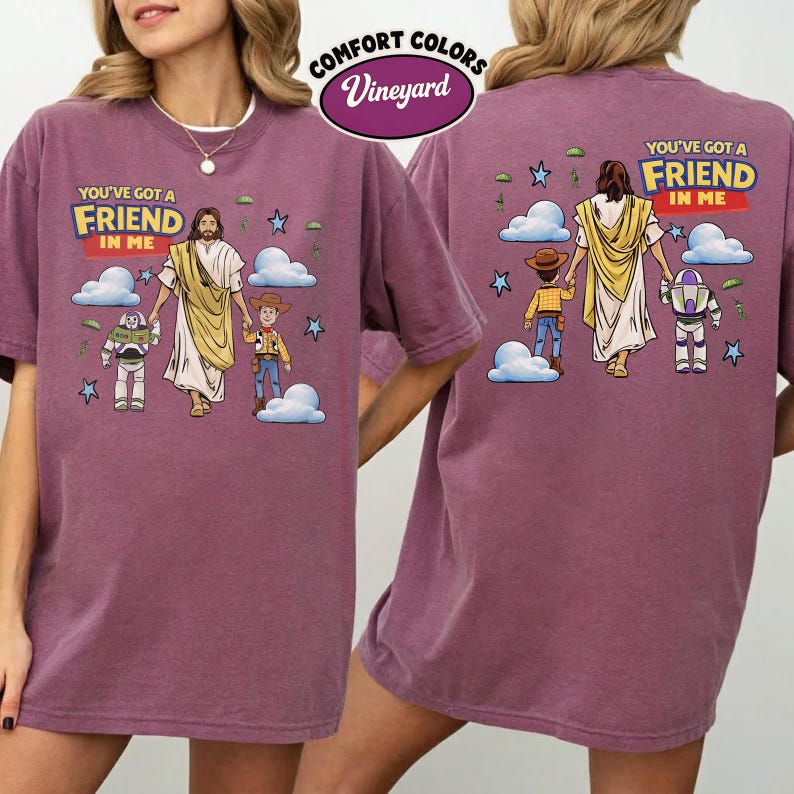 May include: A maroon Comfort Colors t-shirt with a graphic of Jesus and cartoon characters. The text "YOU'VE GOT A FRIEND IN ME" is printed on the front and back. The shirt has a crew neck and short sleeves.