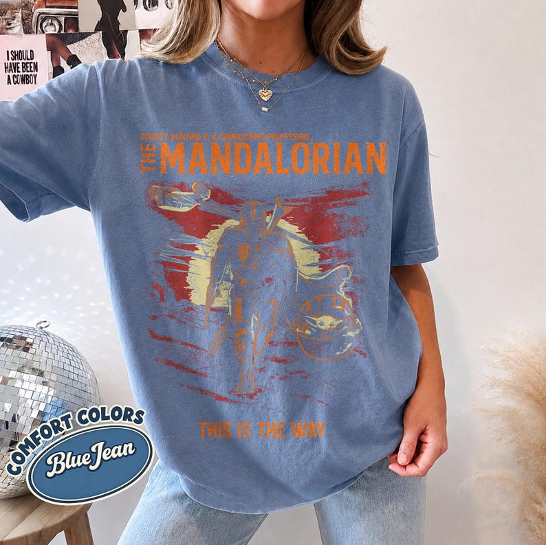 May include: A blue jean colored t-shirt featuring a distressed graphic print of The Mandalorian. The design includes the text The Mandalorian, This Is The Way, and a silhouette of the character against a yellow sun and red background.