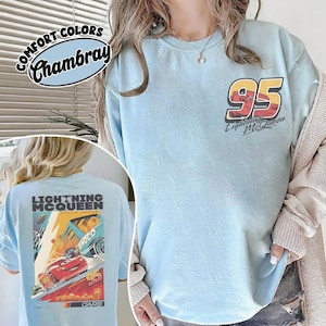 May include: Light blue t-shirt with "95 Lightning McQueen" graphic on the front and a larger graphic on the back. The back graphic features Lightning McQueen racing, with the text "Lightning McQueen" and "Cars". The shirt is labeled "Comfort Colors Chambray".