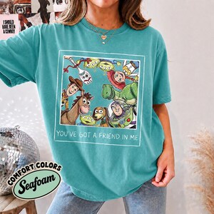 Puede incluir: Camiseta Comfort Colors verde espuma de mar con un gr&aacute;fico enmarcado de personajes de Toy Story. El gr&aacute;fico incluye a Woody, Buzz Lightyear y otros personajes. El texto "YOU'VE GOT A FRIEND IN ME" est&aacute; debajo.