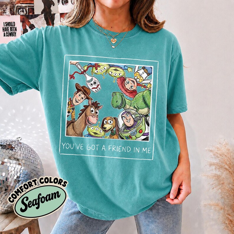Puede incluir: Camiseta Comfort Colors verde espuma de mar con un gr&aacute;fico enmarcado de personajes de Toy Story. El gr&aacute;fico incluye a Woody, Buzz Lightyear y otros personajes. El texto "YOU'VE GOT A FRIEND IN ME" est&aacute; debajo.
