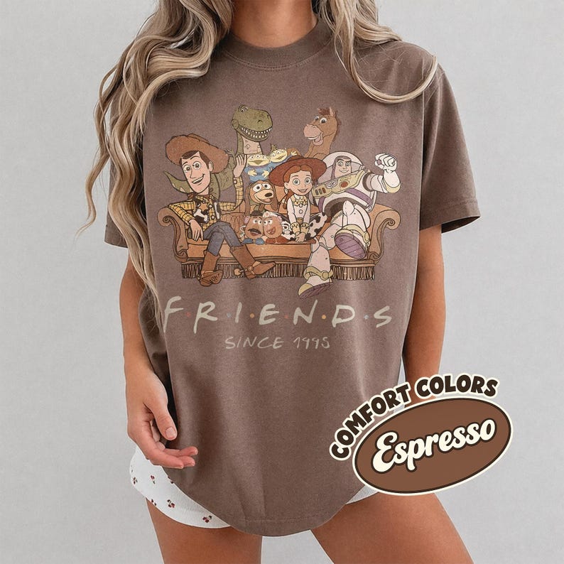 Puede incluir: Camiseta color espresso con un gr&aacute;fico de personajes de Toy Story sentados en un sof&aacute;. La palabra "FRIENDS" est&aacute; encima de "SINCE 1995". La camiseta tiene una etiqueta Comfort Colors.