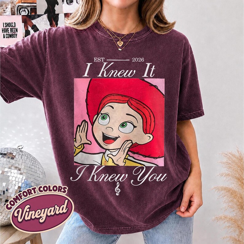 May include: A dark maroon short-sleeve t-shirt featuring a graphic of a red-haired character in a red hat. The text reads EST 2026 I Knew It I Knew You with a small musical note symbol. The shirt has a crew neckline and a vintage-washed appearance.