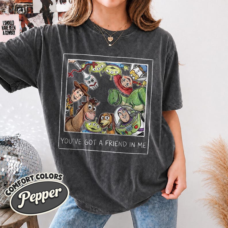 Puede incluir: Camiseta gris oscuro con un gr&aacute;fico enmarcado de personajes de Toy Story. La imagen incluye a Woody, Buzz Lightyear y otros personajes. El texto "YOU'VE GOT A FRIEND IN ME" est&aacute; debajo. La camiseta tiene el logo Comfort Colors Pepper.