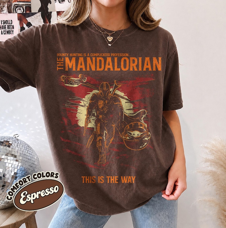 May include: A dark brown short-sleeve t-shirt featuring a graphic of The Mandalorian and Grogu. The text reads: BOUNTY HUNTING IS A COMPLICATED PROFESSION... THE MANDALORIAN and THIS IS THE WAY. The design includes a yellow sun and red accents.