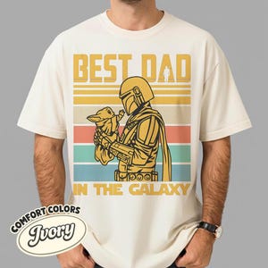 以下が含まれることがあります： アイボリー色のTシャツ。マンダロリアン風のキャラクターが小さな生き物を抱いているグラフィックが特徴です。「BEST DAD IN THE GALAXY」の文字が黄色でプリントされています。シャツにはComfort Colorsのラベルが付いています。