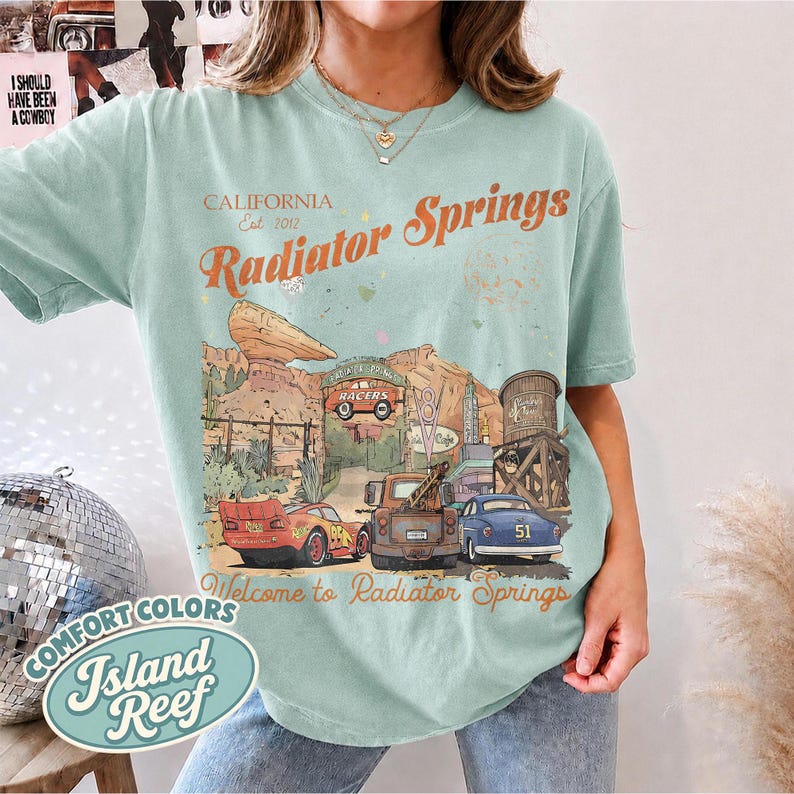 May include: A light green short-sleeve t-shirt featuring a graphic print of Radiator Springs with the text California Est 2012 Radiator Springs and Welcome to Radiator Springs. The illustration includes a red race car, a brown truck, and a blue car.