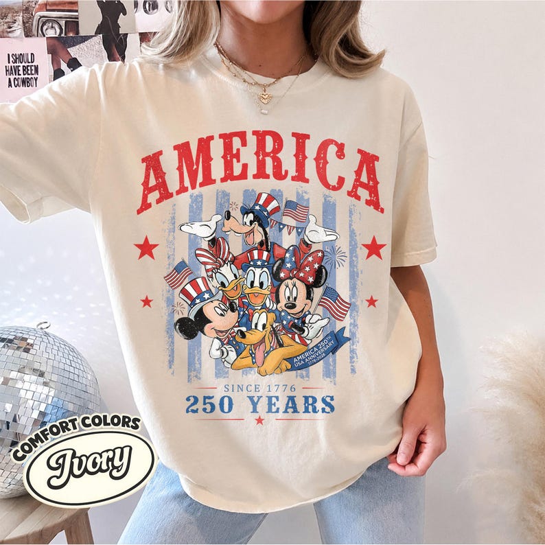 May include: An ivory t-shirt featuring a red, white, and blue graphic of Disney characters wearing patriotic hats. The text reads AMERICA, SINCE 1776, 250 YEARS, and AMERICA 250TH USA ANNIVERSARY. The design includes stars, stripes, and small American flags.